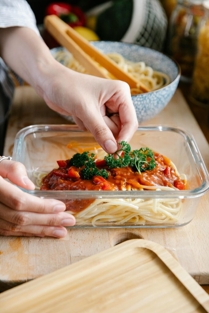 Crop anonymous cook putting green parsley into glass container with pasta and tasty bolognese sauce while cooking at kitchen counter